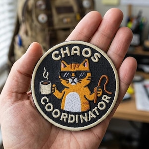 May include: A circular embroidered patch with a black background and a cream-colored border. It features an orange cat wearing black sunglasses, holding a steaming white mug and a brown whip. The text reads CHAOS COORDINATOR in cream letters.