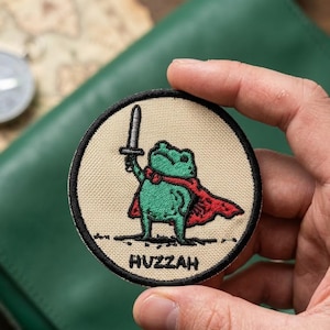 May include: A circular embroidered patch with a beige background and a black border. The design features a green frog wearing a red cape, holding a grey sword aloft. The word HUZZAH is embroidered in black capital letters at the bottom of the patch.