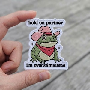 May include: An embroidered patch featuring a green frog wearing a pink cowboy hat and a red bandana. The text "hold on partner" is embroidered in black above the frog, and "i'm overstimulated" is embroidered in black below the frog on a white background.