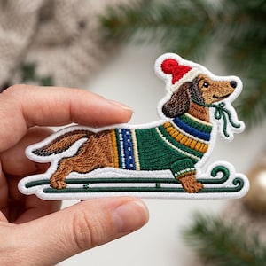 May include: An embroidered patch featuring a brown dachshund wearing a red and white knit hat and a green, blue, and yellow sweater. The dog is depicted riding on a dark green sled and holding a small green string in its mouth.