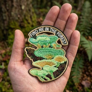 May include: An embroidered patch featuring a cluster of light green and yellow mushrooms with dark brown accents. The patch has a black border and the text "FIND ME IN THE FOREST" arched across the top in white capital letters.