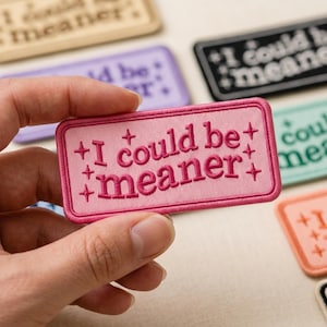 May include: A rectangular pink embroidered patch with a dark pink border and dark pink text that reads "I could be meaner" surrounded by small four-pointed star symbols. The patch is held up against a collection of similar patches in various colors.