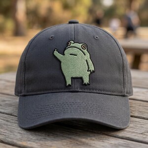 May include: A charcoal gray baseball cap featuring an embroidered patch of a light green frog standing upright. The frog is depicted with a neutral expression and is raising one of its arms, with its middle finger extended outward.
