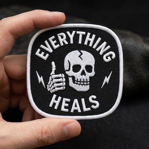 May include: A square embroidered patch with rounded corners featuring a black background and a white border. The design includes a white skull with a cracked forehead, a skeletal hand giving a thumbs up, two lightning bolts, and the text EVERYTHING HEALS.