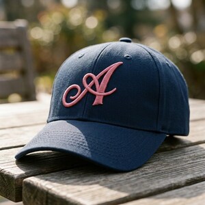 K&ouml;nnte beinhalten: Eine marineblaue Baseballkappe mit einem markanten, gestickten Buchstaben A in einer hellrosa Schreibschrift. Die Kappe verf&uuml;gt &uuml;ber eine strukturierte Krone, einen gebogenen Schirm und gen&auml;hte &Ouml;sen an den oberen Paneelen.