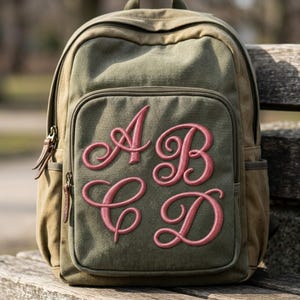 K&ouml;nnte beinhalten: Ein olivgr&uuml;ner Rucksack aus Segeltuch mit einer gro&szlig;en Rei&szlig;verschlusstasche auf der Vorderseite. Die Vordertasche ist mit den Buchstaben A, B, C und D in einer gro&szlig;en, rosafarbenen Schreibschrift bestickt. Der Rucksack hat braune Lederzipper.