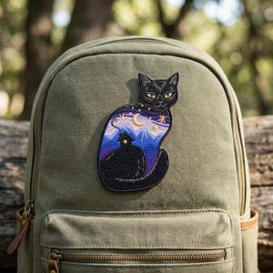 May include: An olive green canvas backpack featuring an embroidered patch of a black cat. The cat's body contains a landscape scene with a small house, blue mountains, a purple night sky, a yellow crescent moon, and several golden stars.