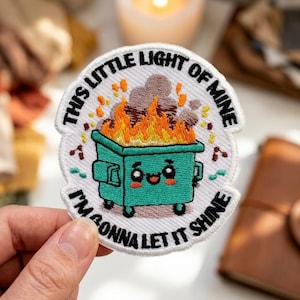May include: A circular embroidered patch featuring a teal dumpster with a smiling face, filled with orange and yellow flames and grey smoke. The text reads THIS LITTLE LIGHT OF MINE and I'M GONNA LET IT SHINE in black capital letters.