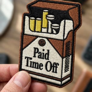 May include: An embroidered patch shaped like a cigarette pack with a brown and white color scheme. The design features yellow-tipped cigarettes inside the pack and the black text Paid Time Off printed on the front in a bold serif font.