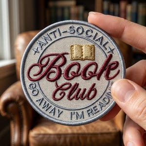 May include: A circular embroidered patch with a beige fabric background and a grey border. It features the text "ANTI-SOCIAL BOOK CLUB" and "GO AWAY I'M READING" in grey, with the words "Book Club" in dark red script and a small gold book icon.