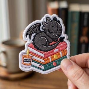 May include: An embroidered patch featuring a small black dragon sleeping on a stack of three books in red, orange, and green. A small orange candle with a yellow flame sits beside the books. The entire design is outlined in white thread.