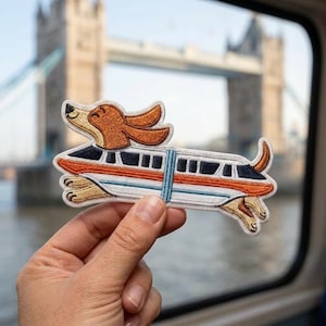 May include: An embroidered patch featuring a brown and tan dog with long ears integrated into the body of a white, orange, and blue train car. The dog's head and paws extend from the train, which has black windows and a blue vertical stripe.