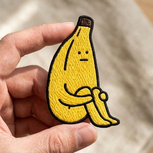 May include: A yellow banana-shaped embroidered patch with a black border. The banana has a brown stem, a simple face with two dots for eyes and a straight line for a mouth, and is depicted in a seated position with arms wrapped around its knees.