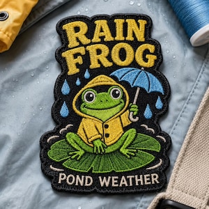 May include: An embroidered patch featuring a green frog wearing a yellow raincoat and holding a blue umbrella while sitting on a lily pad. The text RAIN FROG appears in yellow at the top, and POND WEATHER is written in white at the bottom.