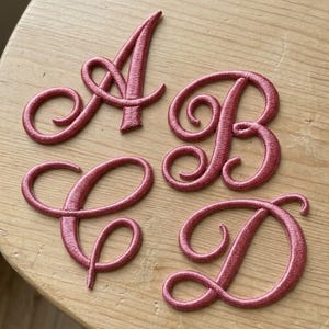 K&ouml;nnte beinhalten: Vier einzelne gestickte Buchstabenaufn&auml;her in Altrosa, die die Gro&szlig;buchstaben A, B, C und D in einer kursiven Schriftart mit Satin-Stich-Finish und einer erhabenen, strukturierten Optik darstellen.