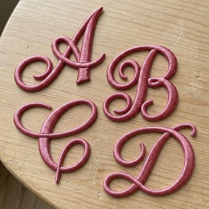 K&ouml;nnte beinhalten: Vier einzelne gestickte Buchstabenaufn&auml;her in Altrosa, die die Gro&szlig;buchstaben A, B, C und D in einer kursiven Schriftart mit Satin-Stich-Finish und einer erhabenen, strukturierten Optik darstellen.