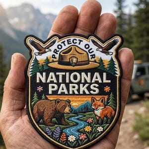 May include: A shield-shaped embroidered patch featuring a brown bear and a red fox near a blue river. The design includes a tan ranger hat, two eagles, green pine trees, and colorful flowers. The text reads PROTECT OUR NATIONAL PARKS in white.