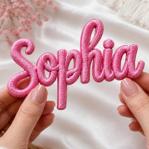May include: A bright pink embroidered patch featuring the name Sophia in a cursive script font. The lettering has a raised, textured appearance with a metallic sheen, showcasing dense stitching throughout the entire surface of the name design.