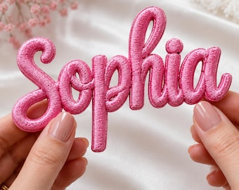 May include: A bright pink embroidered patch featuring the name Sophia in a cursive script font. The lettering has a raised, textured appearance with a metallic sheen, showcasing dense stitching throughout the entire surface of the name design.