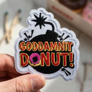 May include: A black embroidered patch shaped like a bomb with a lit fuse. The text reads GODDAMNIT DONUT! in red and yellow letters, with a pink and yellow donut replacing the letter O. The patch has a white border and a white stitched background.