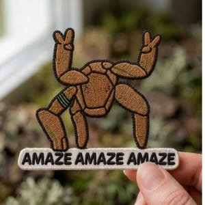 May include: An embroidered patch featuring a brown, multi-legged creature with green accents, standing on a white rectangular base. The text "AMAZE AMAZE AMAZE" is stitched in black capital letters across the base of the patch.