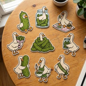 May include: A collection of nine embroidered patches featuring white ducks interacting with green pickles. Designs include a duck in a pickle suit, a duck in a jar, a duck on a picnic, a duck holding a pickle bouquet, a duck in a pickle pile, a duck carrying a jar, a duck hugging a pickle, a duck cooking, and a duck wearing a Pickle King sash.