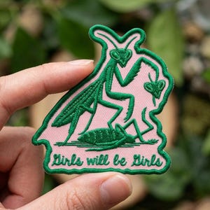 May include: An embroidered patch featuring a green praying mantis on a light pink background. The design shows a mantis standing over another insect with the text Girls will be Girls stitched in green thread at the bottom of the patch.