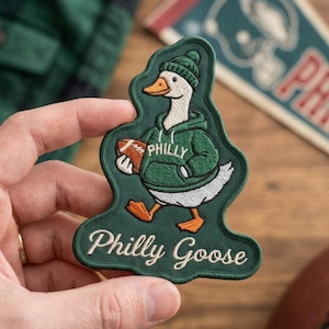 May include: An embroidered patch featuring a white goose wearing a green hoodie and a matching green beanie. The goose holds a brown football. The hoodie has the word Philly printed on it. The text Philly Goose is embroidered at the bottom.