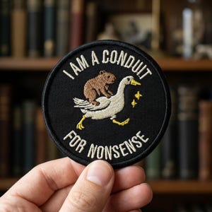 May include: A circular black embroidered patch featuring a white goose carrying a brown capybara on its back. The text "I AM A CONDUIT FOR NONSENSE" is stitched in white thread around the top and bottom edges, with small yellow stars nearby.