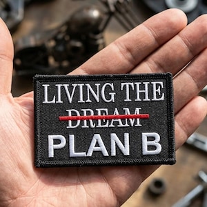 May include: A rectangular black embroidered patch with white text that reads LIVING THE, followed by the word DREAM with a horizontal red line struck through it, and the text PLAN B underneath. The patch has a black stitched border.