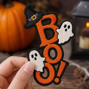 May include: An embroidered patch featuring the orange text BOO! with a black outline. The design includes a black witch hat with a purple band and yellow buckle atop the letter B, and two small white ghosts with black eyes and mouths.