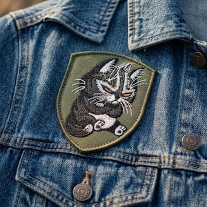 May include: A shield-shaped embroidered patch featuring a grumpy black and white cat with orange eyes, stitched onto a blue denim jacket. The patch has an olive green background and a dark green border, showcasing detailed thread work of the cat.