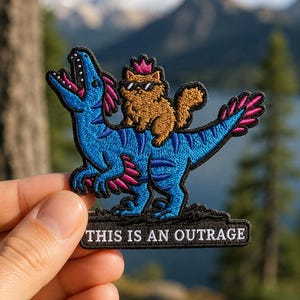 May include: An embroidered patch featuring a blue dinosaur with pink accents and an open mouth, carrying a brown cat wearing pink sunglasses and a pink crown. Below the figures, a black rectangular banner displays the text THIS IS AN OUTRAGE.