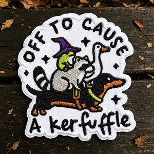 May include: A white embroidered patch featuring a black dachshund, a gray raccoon, a white goose, and a green frog wearing a purple witch hat. The text "OFF TO CAUSE A KERFUFFLE" is stitched in black around the animals, accented by small black stars.