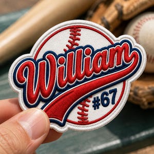 May include: A circular embroidered patch featuring a white baseball design with red stitching. The name William is stitched in bold red script with a dark blue outline. Below the name, the text #67 is embroidered in dark blue on a red swoosh.