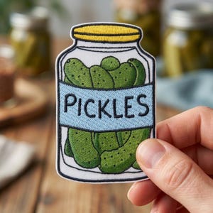 May include: An embroidered patch shaped like a glass jar filled with green pickles. The jar has a yellow lid and a light blue label across the center with the word PICKLES stitched in black thread. The design features a black border outline.