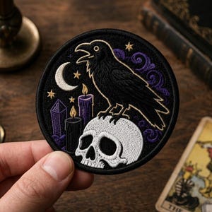 May include: A circular embroidered patch featuring a black raven perched on a white human skull. The design includes a white crescent moon, gold stars, two purple candles with yellow flames, and purple swirling clouds on a black background.