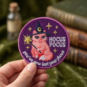 May include: A circular purple embroidered patch featuring a pink cat wearing a black witch hat with a yellow crescent moon. The cat holds a yellow star wand. Text reads HOCUS POCUS and you've now lost your focus, surrounded by yellow stars.