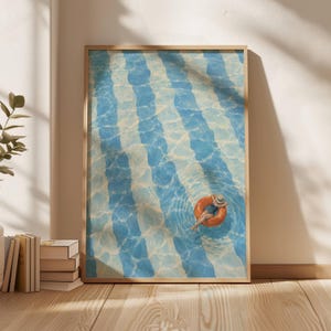 Könnte beinhalten: Ein gerahmter vertikaler Wandkunstdruck mit einer Luftaufnahme eines Schwimmbeckens mit blauen und weißen vertikalen Streifen. Eine Person mit Hut und Badeanzug treibt auf einem leuchtend orangefarbenen, runden Schwimmring.