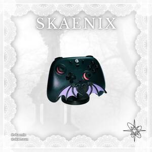 May include: A black video game controller featuring a central attachment shaped like a dark heart with light purple bat wings. The controller has red accents on the analog sticks and the text SKAENIX is displayed in white at the top of the image.