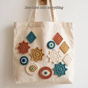 May include: A beige canvas tote bag decorated with various colorful crochet appliques, including star shapes, diamonds, and circular evil eye motifs. The text "Sew them onto everything" is printed in black above the bag handles.
