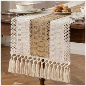 May include: A rectangular table runner featuring a central tan woven panel flanked by cream-colored sections with intricate crochet patterns and openwork borders. The ends are finished with long, cream-colored knotted fringe tassels.