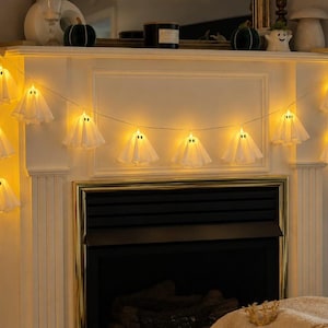 May include: A string of LED fairy lights featuring multiple white fabric ghost covers, each with two small black eyes. The lights emit a warm yellow glow and are draped across a fireplace mantel, creating a festive illumination for seasonal decor.
