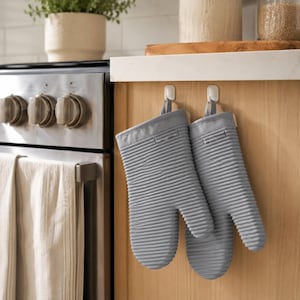 May include: A pair of light gray oven mitts with a horizontal ribbed texture hanging from white adhesive hooks. The mitts feature a thumb section and a main hand compartment, constructed with a consistent ridged pattern across the entire surface.