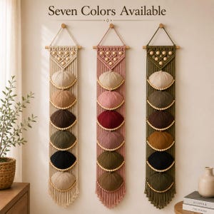 May include: Three vertical macrame wall hangings with wooden dowels and hanging loops. Each piece features a geometric pattern with wooden beads and six circular pockets holding baseball caps in various colors. Text reads Seven Colors Available.