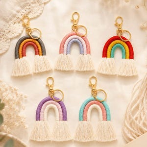 May include: Five macrame rainbow keychains featuring gold-tone lobster clasps and rings. Each keychain displays a unique color combination of wrapped cord arches ending in cream-colored fringe tassels. The designs include grey, orange, pink, blue, and purple.
