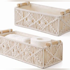 May include: Two rectangular storage baskets featuring an off-white macrame exterior with intricate knotted patterns, a solid off-white fabric lining, and two light-colored wooden handles on the sides. One basket contains a white towel and toilet paper.