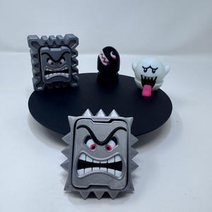 Op de afbeelding: Een verzameling van vier kleine plastic figuren met twee grijze stekelige Thwomp-blokken met boze gezichten, een zwarte Bullet Bill met witte tanden en een witte Boo-geest met een roze tong, geplaatst op een ronde zwarte basis.