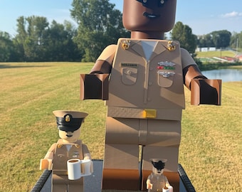 May include: A large, oversized plastic figure dressed in a tan United States Navy Chief Petty Officer uniform, featuring a black peaked cap with a gold emblem, a name tag reading BENJAMIN, and various service pins. Two smaller matching figures stand nearby.