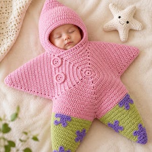 May include: A pink crocheted baby swaddle shaped like a star with a hood, featuring two round buttons on the chest and light green lower sections decorated with purple flower-like shapes, accompanied by a small cream-colored crocheted starfish toy.